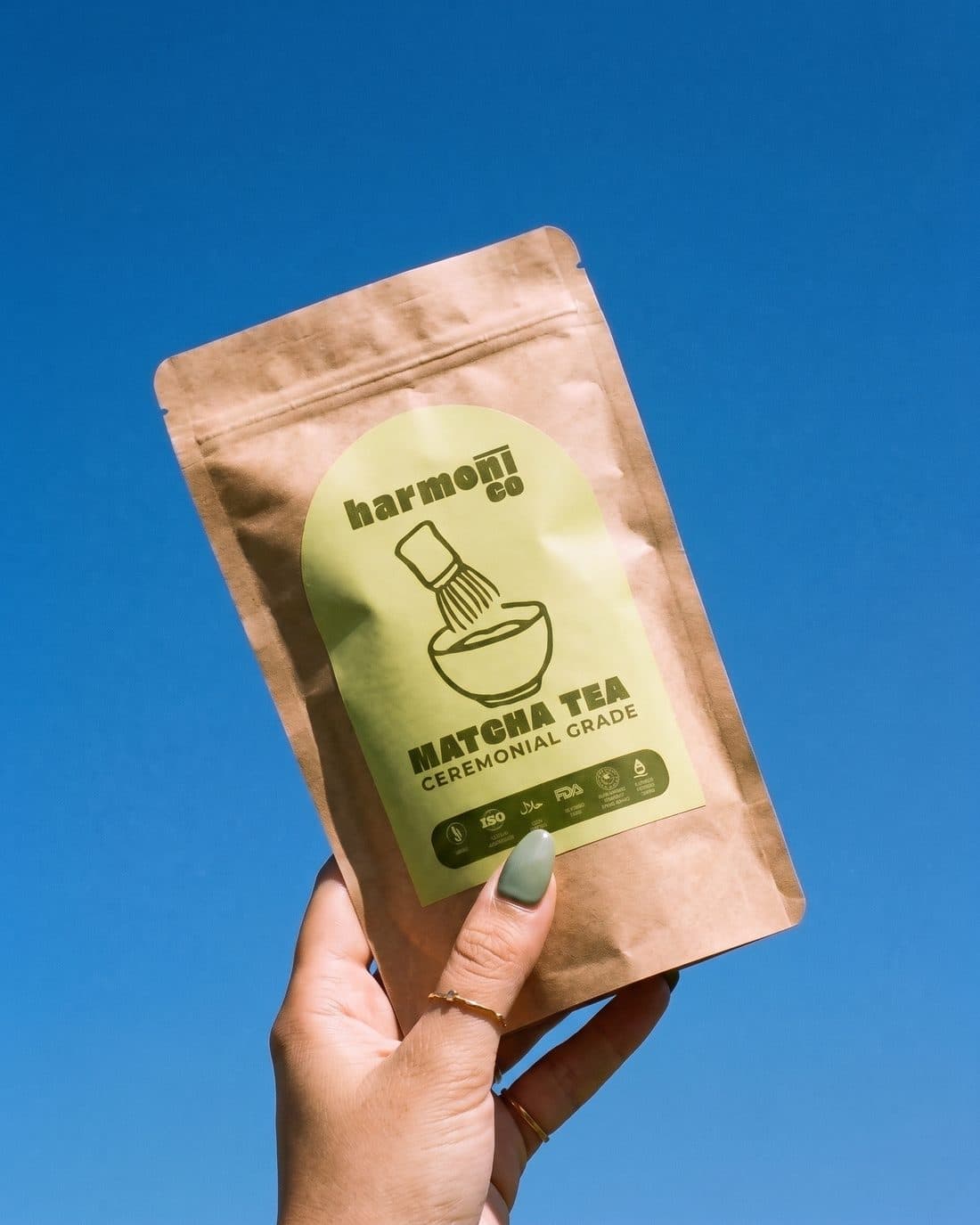 Adaçayı ojeli bir el Harmonico matcha kesesini mavi gökyüzüne kaldırıyor